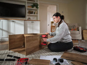 Person assembling wooden furniture on a carpeted floor using a red CRAFTSMAN cordless drill, with toolboxes, hardware, and instructions spread nearby in a living room setting.
