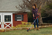 Photo of a person using a Craftsman cordless string trimmer, SKU CMCK397M1, trimming grass near a red shed outdoors.