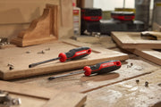 Two CRAFTSMAN screwdrivers with black and red handles are placed on a wooden workbench surrounded by wood pieces and metal screws. The workspace appears to be a carpentry or workshop setting.