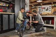 A person kneeling in a workshop uses a hand tool to repair a blue bicycle while another person stands nearby. The workshop