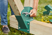 A person is tightening a bolt with a ratchet wrench on a green metal connector that joins wooden beams outdoors, possibly as part of a construction or assembly project.