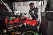 A person organizing red and black Craftsman toolboxes and a tool bag in the back of a vehicle, with various tools and equipment visible, suggesting a well-equipped mobile workshop.