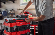 A person is organizing a stack of red and black CRAFTSMAN toolboxes in a garage or workshop setting. The toolboxes are part of a modular storage system and have handles and latches for secure stacking.