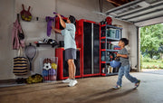 A person is organizing sports equipment in a garage with red and black CRAFTSMAN storage cabinets and shelves. A child is holding a bicycle helmet and walking towards the adult. Various sports balls, tennis rackets, and bags are visible, along with a large basket for balls and open garage door with greenery outside.