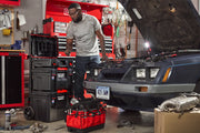 A person working in a garage is using various Craftsman tool storage boxes and a red tool tote, with a car parked nearby that has its hood open for maintenance. Several tools and automotive parts are scattered around the workspace. Shelves and tool cabinets are visible in the background.