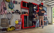 A garage scene featuring red and black CRAFTSMAN storage cabinets (SKU: CMST82602VT) filled with various sport equipment, toys, and supplies. Two people are organizing items in the cabinets. Shelves and baskets containing balls, rackets, and other gear are visible throughout the space.