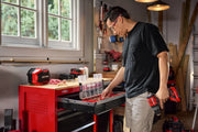 A person standing in a workshop is organizing tools in a red Craftsman tool chest. The individual, whose face is blurred, holds a red power drill in one hand and sorts through a drawer filled with various toolkit items. The workspace includes tool storage, pegboards with tools, and windows letting in natural light.