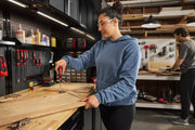 Two people are working in a workshop. One person in the foreground is using a CRAFTSMAN screwdriver to assemble wooden boards on a workbench, while another person is working at a table in the background. The workshop contains various tools and supplies organized on shelves and pegboards.