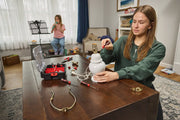 A person is repairing a lamp on a wooden table using hand tools, with a CRAFTSMAN toolbox and several screwdrivers and lamp parts spread out. In the background, another person sits near a window playing a wind instrument. The room appears to be a living space with bookshelves, furniture, and daylight coming through large windows.
