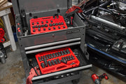 A CRAFTSMAN tool chest with two open drawers containing red trays filled with socket sets and other tools, situated in a garage beside an open car hood. Mechanic gloves and various tools are visible around the chest.