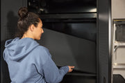A person wearing a blue hoodie is placing or adjusting a large, black shelf inside a metal storage cabinet. Their face is