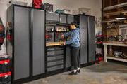 A person is working at a wooden workbench, which is part of a CRAFTSMAN modular storage cabinet system. The cabinet set includes tall cabinets, upper and lower storage units, and a pegboard for organizing tools. Various tools and supplies are visible on and around the workbench in a garage workshop setting.